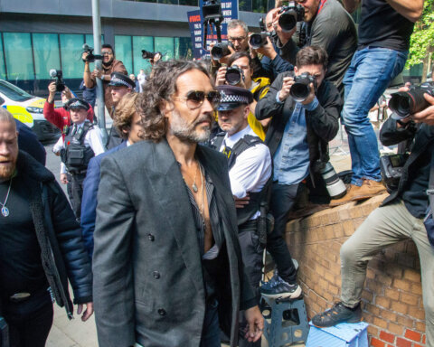 Russell Brand wearing sunglasses and a dark blazer walking past photographers and police officers.