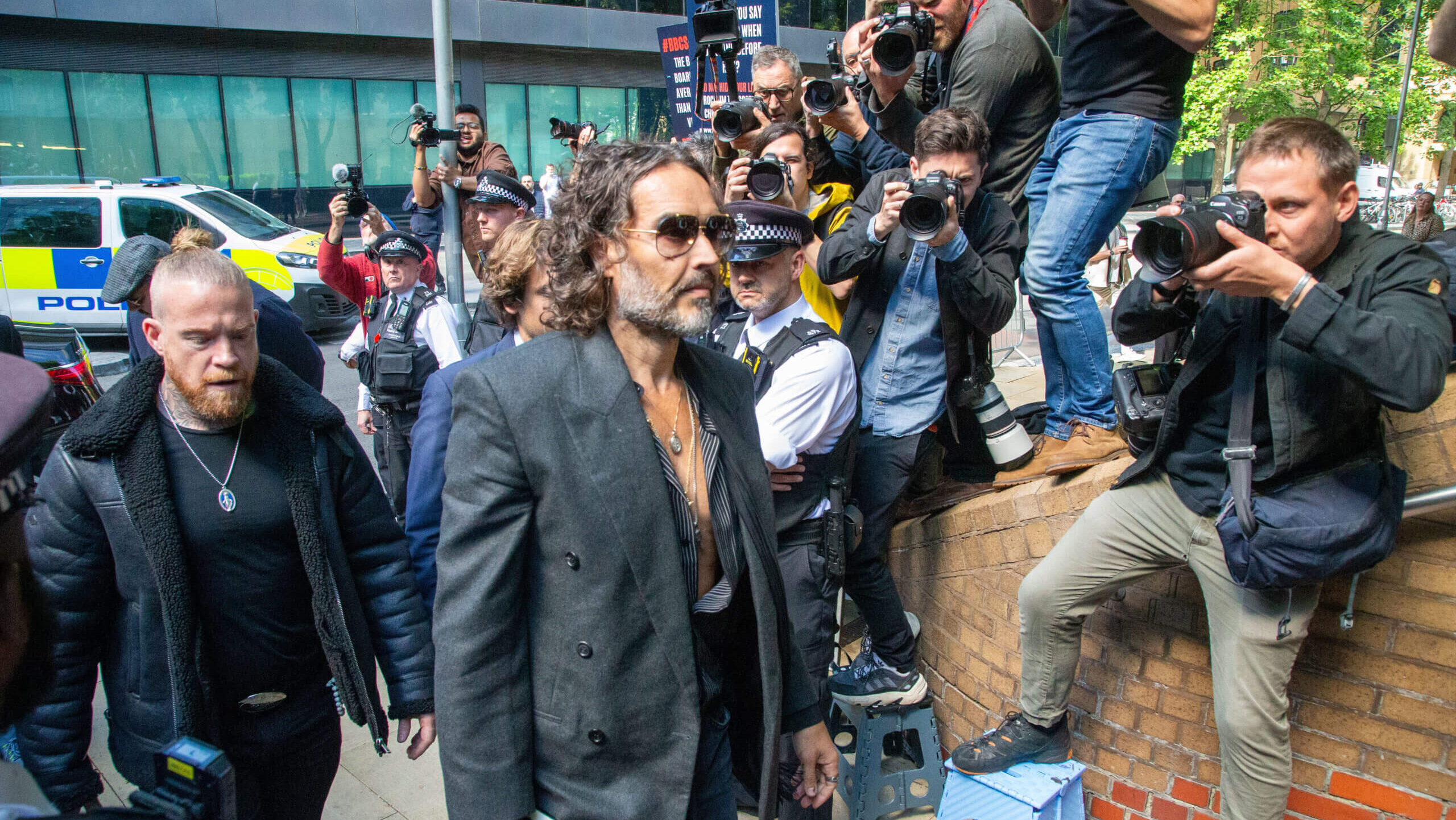 Russell Brand wearing sunglasses and a dark blazer walking past photographers and police officers.