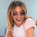 Smiling woman with blonde hair wearing round sunglasses and a light pink t-shirt