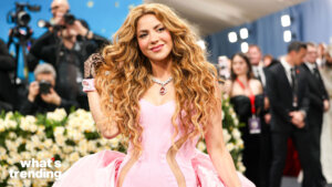 Shakira in a pink gown with long curly hair posing on a red carpet with photographers in the background
