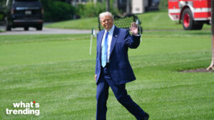 Donald Trump walking on grass waving, wearing a navy suit and light blue tie