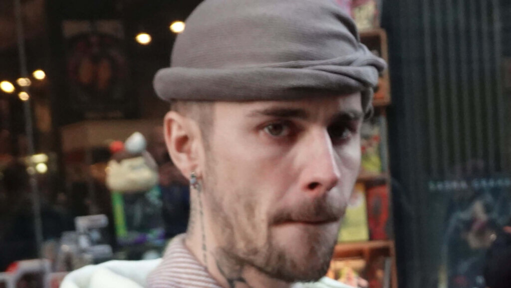 man wearing a gray beanie and striped shirt with ear and neck tattoos in an indoor setting