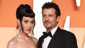Katy Perry and Orlando Bloom posing together at a formal event with an orange and white background.