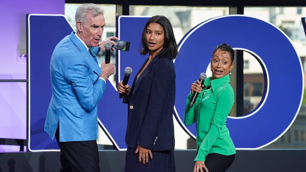 Bill Nye holding a phone and microphone with two women holding microphones in front of large blue letters.