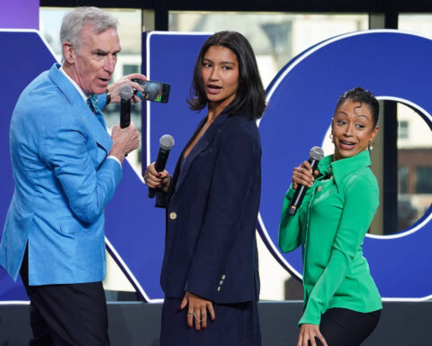 Bill Nye holding a phone and microphone with two women holding microphones in front of large blue letters.