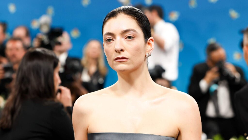 Lorde with slicked-back hair and strapless black dress at a formal event with photographers in the background