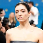 Lorde with slicked-back hair and strapless black dress at a formal event with photographers in the background