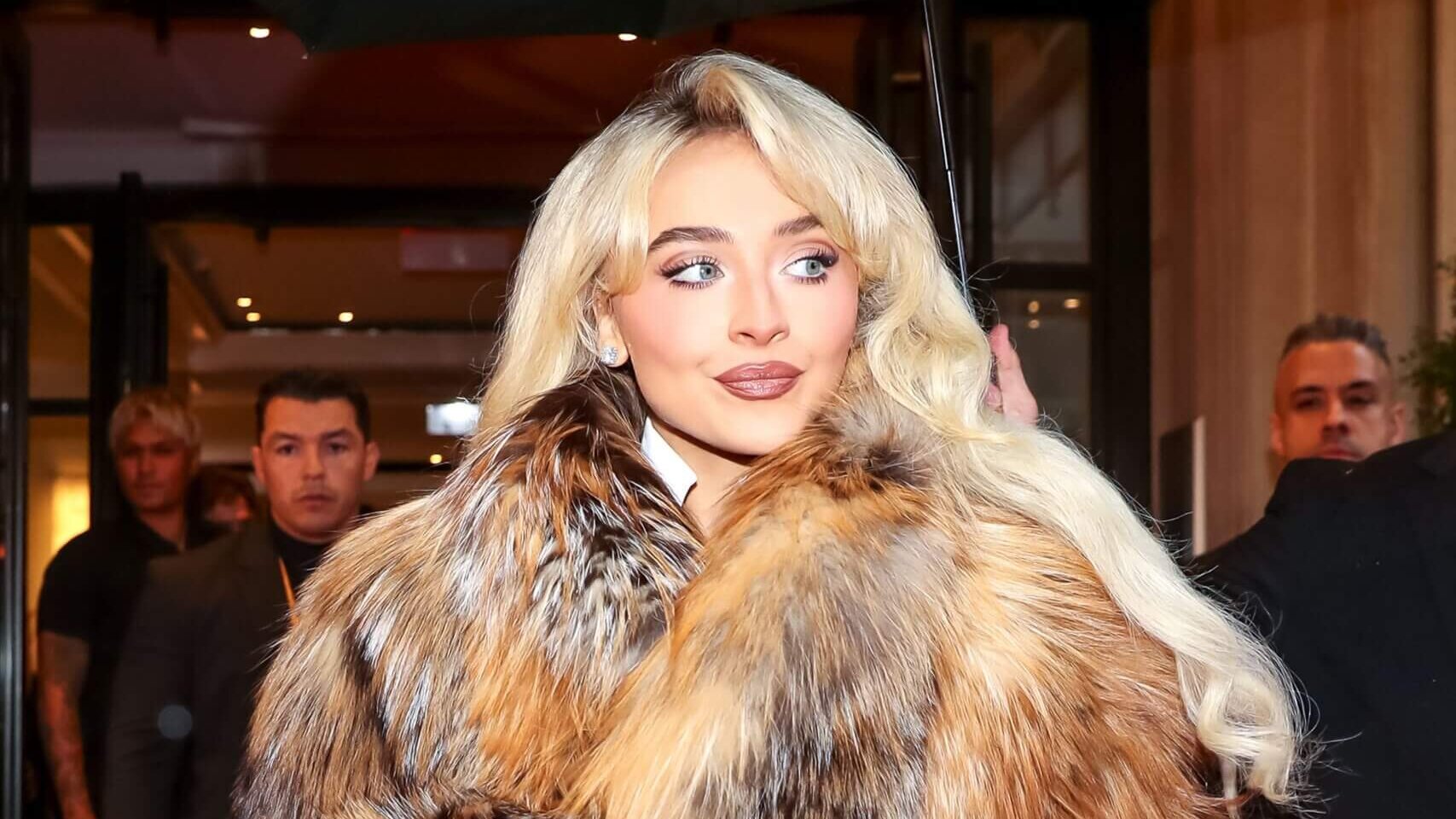 Blonde woman wearing a large fur coat looking to the side with people in the background indoors