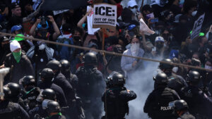 Protesters holding "ICE: Out of Our Communities!" signs face police officers in riot gear amid smoke.