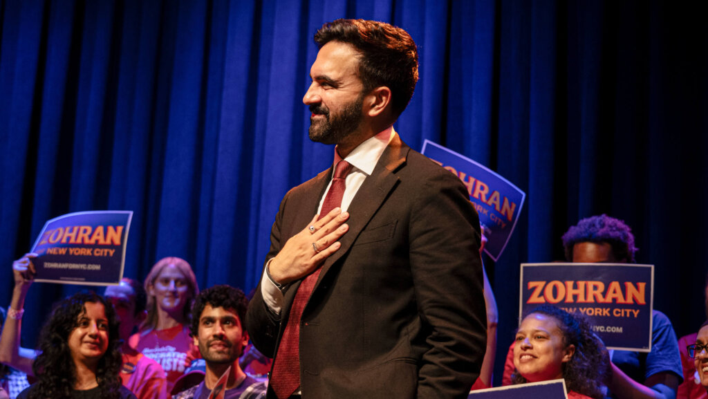 Man in suit and red tie with hand on chest at a campaign event with supporters holding "Zohran New York City" signs