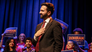 Man in suit and red tie with hand on chest at a campaign event with supporters holding "Zohran New York City" signs