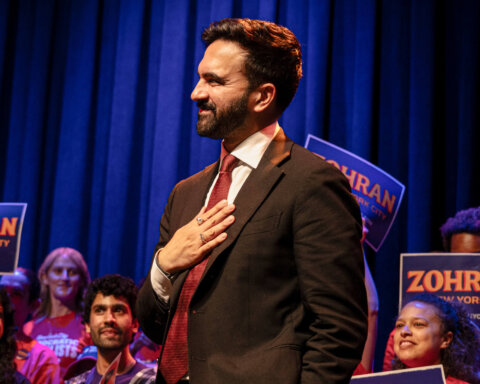 Man in suit and red tie with hand on chest at a campaign event with supporters holding "Zohran New York City" signs