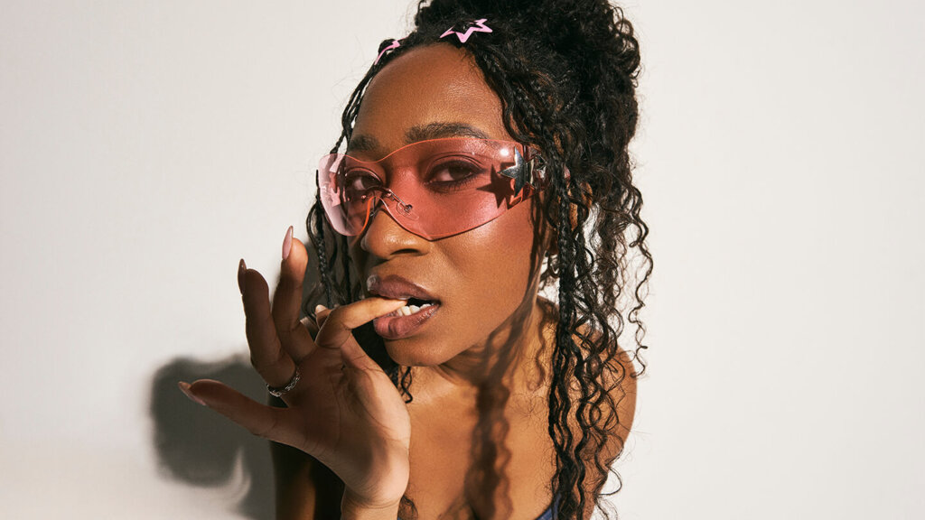 Woman with curly hair wearing pink star-shaped sunglasses and hair clips, biting her finger against a white background