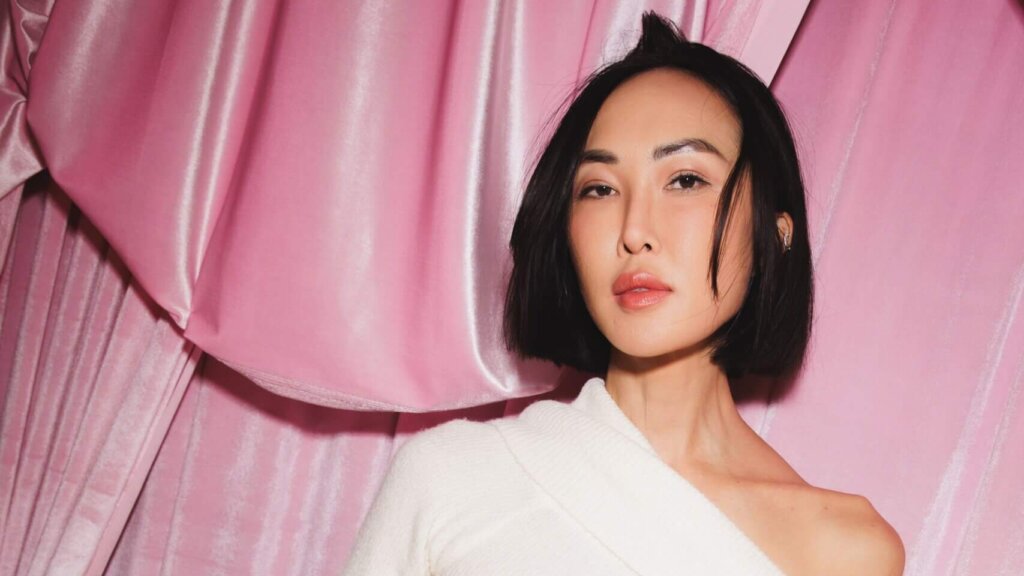 Woman with short black hair wearing a white off-shoulder top against a pink satin curtain background