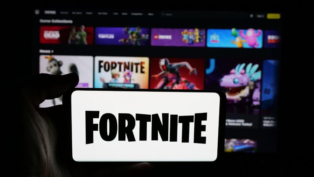 Hand holding smartphone displaying Fortnite logo with blurred gaming platform interface in the background