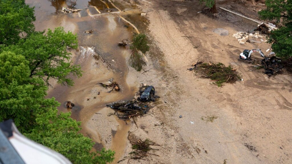 Aerial view of flood debris including a damaged vehicle, uprooted trees, and an excavator on a muddy riverbank.