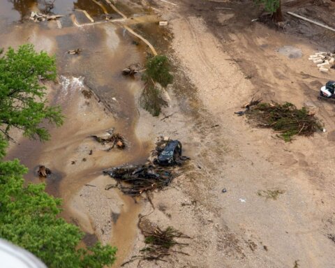 Aerial view of flood debris including a damaged vehicle, uprooted trees, and an excavator on a muddy riverbank.