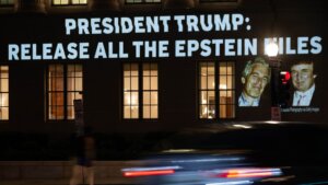 Projection on building reading "President Trump: Release all the Epstein files" with photos of Jeffrey Epstein and Donald Trump at night