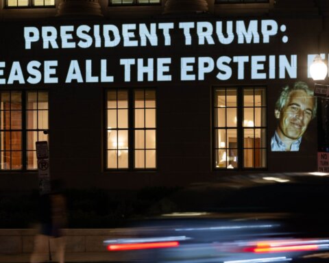 Projection on building reading "President Trump: Release all the Epstein files" with photos of Jeffrey Epstein and Donald Trump at night