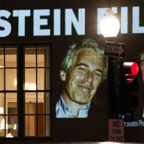 Projection on building reading "President Trump: Release all the Epstein files" with photos of Jeffrey Epstein and Donald Trump at night
