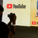 Person taking a photo of a screen displaying the YouTube logo and part of two people wearing shorts and sneakers