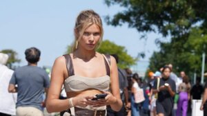 Blonde woman in beige crop top and brown straps holding a phone outdoors with people walking in the background