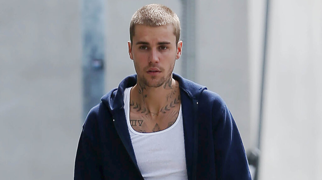 Justin Bieber with short hair, wearing a white tank top and navy hoodie, walking outdoors.