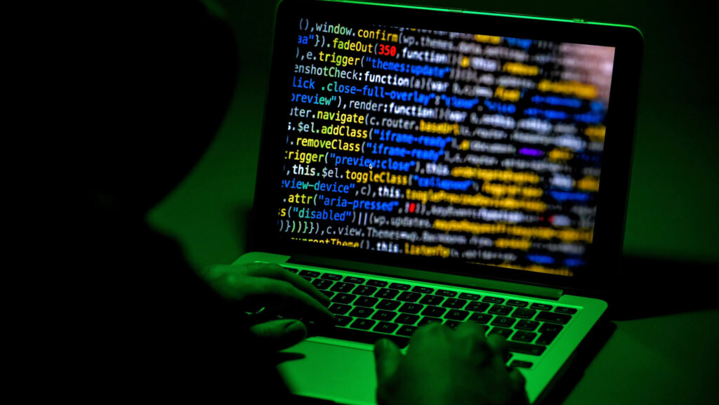 Person typing on a laptop displaying colorful programming code on the screen in a dark environment with green lighting.