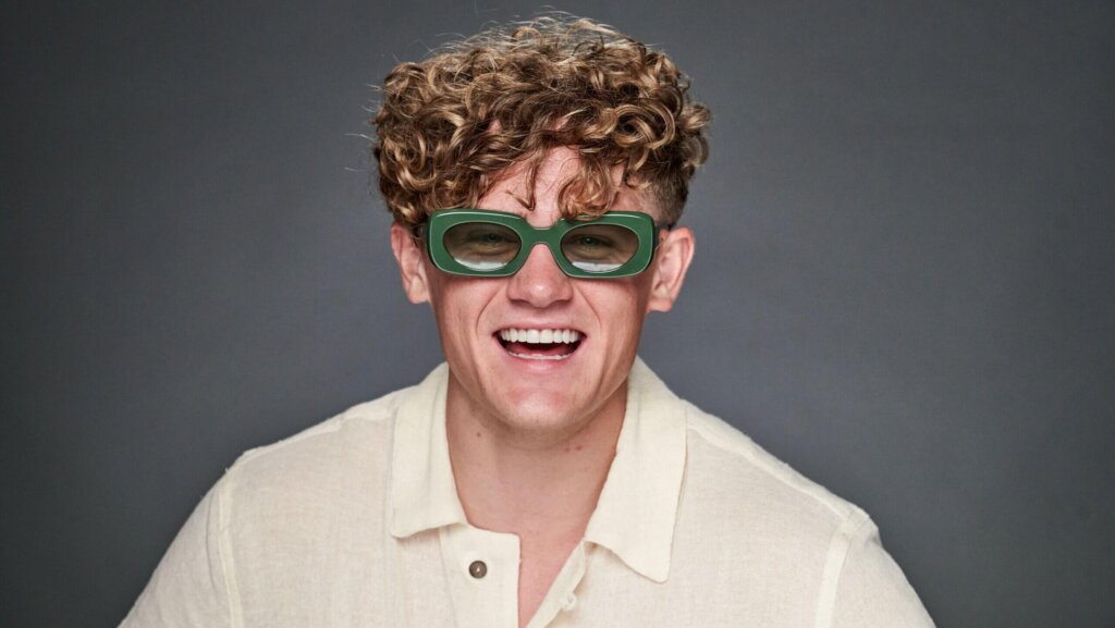 Young man with curly hair wearing green rectangular sunglasses and a cream-colored shirt smiling against a gray background.