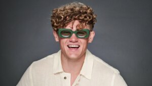 Young man with curly hair wearing green rectangular sunglasses and a cream-colored shirt smiling against a gray background.