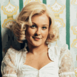 Young woman with blonde curly hair wearing a white lace blouse against a patterned wallpaper background