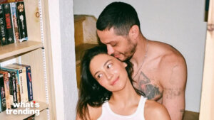 A shirtless man with tattoos embraces a smiling woman in a white tank top near a bookshelf.