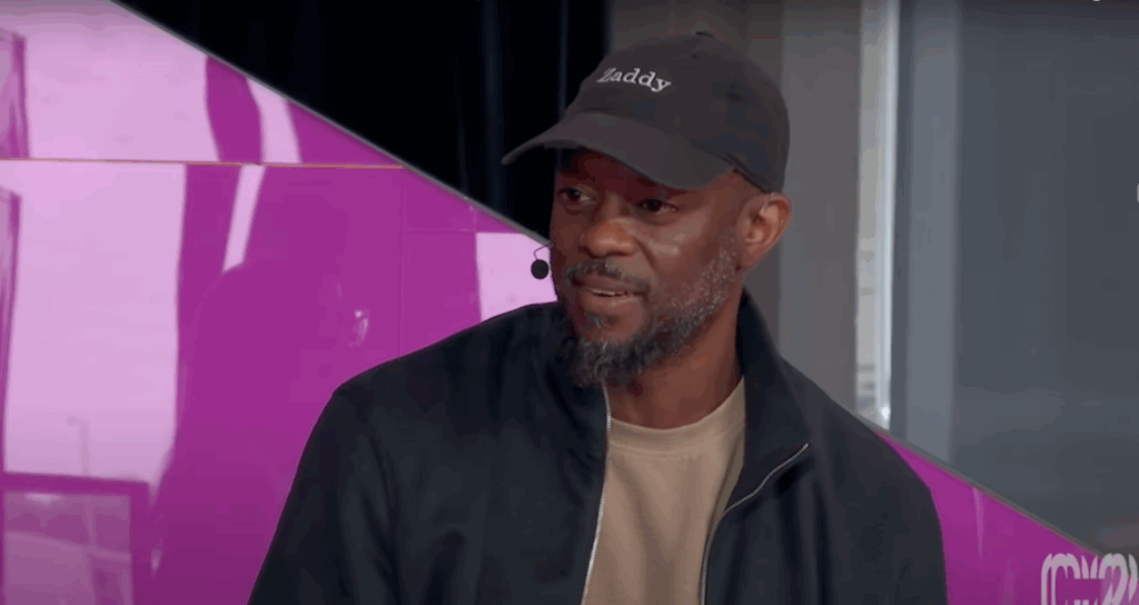 Man wearing a black "Daddy" cap and black jacket speaking with a microphone headset against a purple background