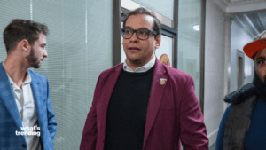 Man in glasses wearing a maroon blazer and black sweater walking in a hallway with two other men