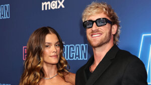 Logan Paul wearing black sunglasses and a black suit posing with a woman with wavy brown hair at a premiere event.