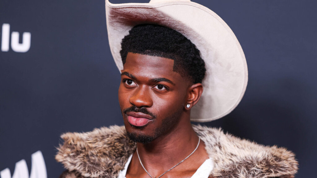 Man wearing a white cowboy hat, fur-collared jacket, and diamond stud earring against a dark background
