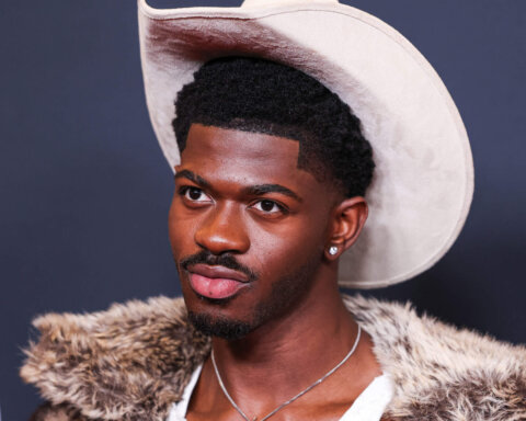 Man wearing a white cowboy hat, fur-collared jacket, and diamond stud earring against a dark background
