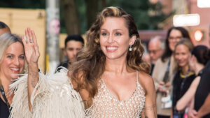 Miley Cyrus smiling and waving, wearing a white fringed outfit with a crowd in the background.
