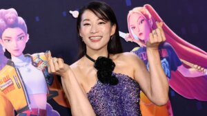 Smiling woman in a purple sequined dress with black rose necklace making finger heart gestures at an event with animated characters backdrop.