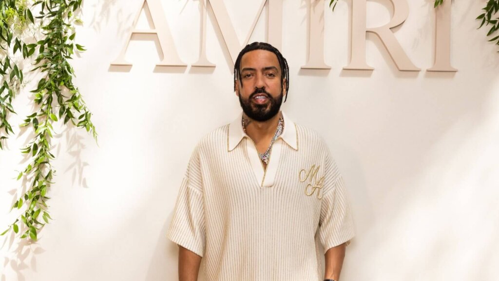 Man with braided hair and beard wearing a cream ribbed shirt with "MH" initials standing in front of "AMIRI" sign