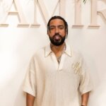 Man with braided hair and beard wearing a cream ribbed shirt with "MH" initials standing in front of "AMIRI" sign