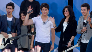 Joe Jonas, Nick Jonas, Demi Lovato, and Kevin Jonas performing on stage with instruments and microphones.