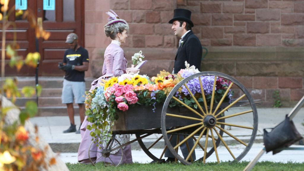 Two people in Victorian attire stand beside a wooden cart filled with colorful flowers on a city sidewalk.