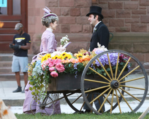 Two people in Victorian attire stand beside a wooden cart filled with colorful flowers on a city sidewalk.