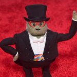 Mr. Potato Head dressed in a tuxedo, top hat, and red heart-shaped glasses on a red carpet background