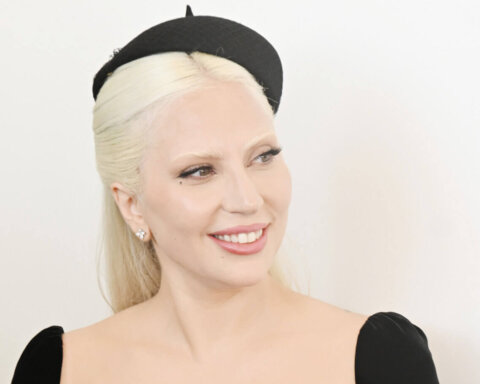 Lady Gaga smiling wearing a black dress and a small black hat against a white background