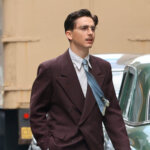 Timothée Chalamet walking on a city street wearing a brown suit and blue tie.