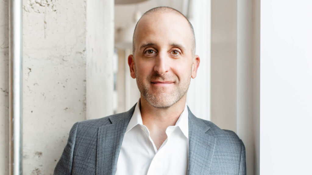 Smiling bald man in a gray suit jacket and white shirt standing by a window and concrete wall