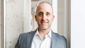 Smiling bald man in a gray suit jacket and white shirt standing by a window and concrete wall