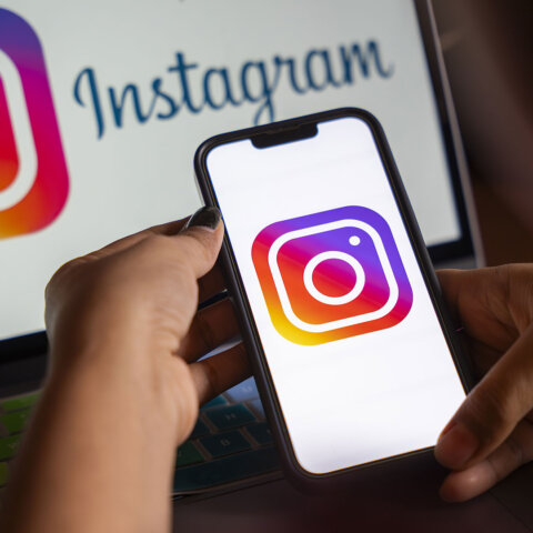 Hands holding a smartphone displaying the Instagram logo with a laptop showing the Instagram logo and text in the background
